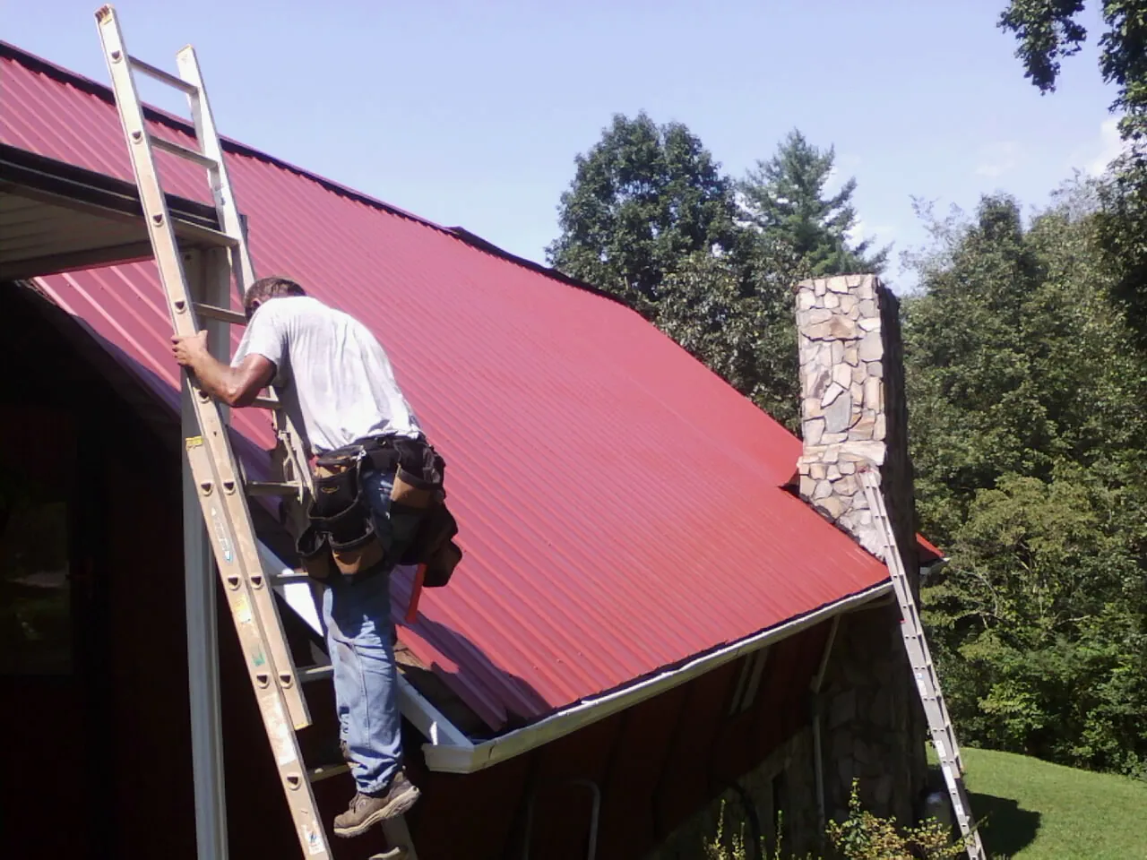 Roofer on a ladder beside a red metal roof and stone chimney — Soffit & Fascia Repair by Roofing Brothers in Sweeden